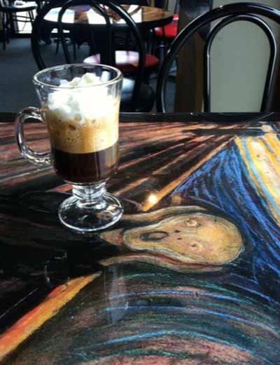 A glass of coffee with whipped cream is on a table next to a painting of a scream.
