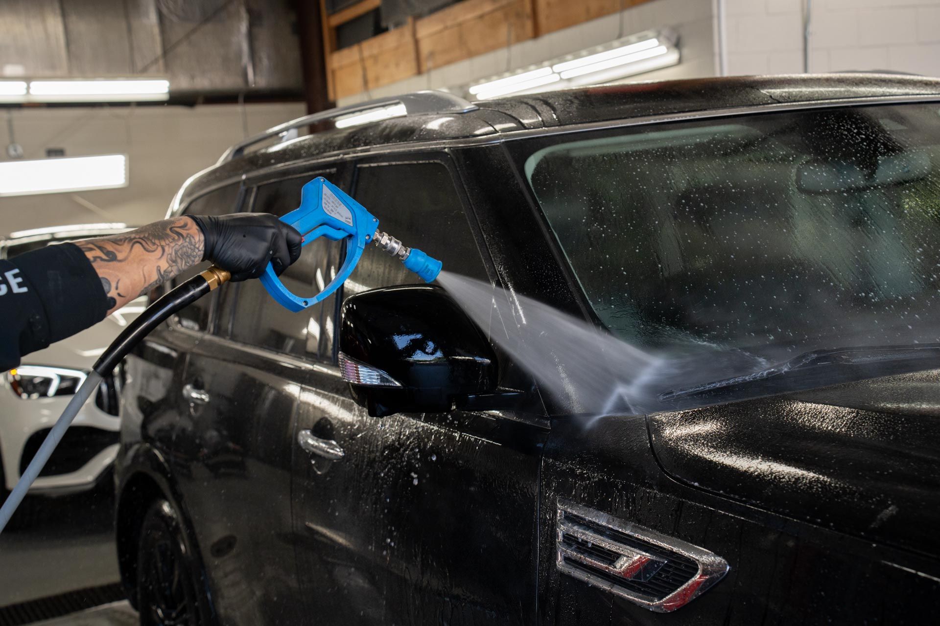 Person washing a black SUV with a pressure washer in an indoor garage setting.