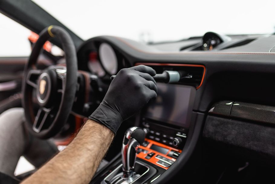 Gloved hand cleaning a car dashboard. A black brush cleans the dashboard's screen.