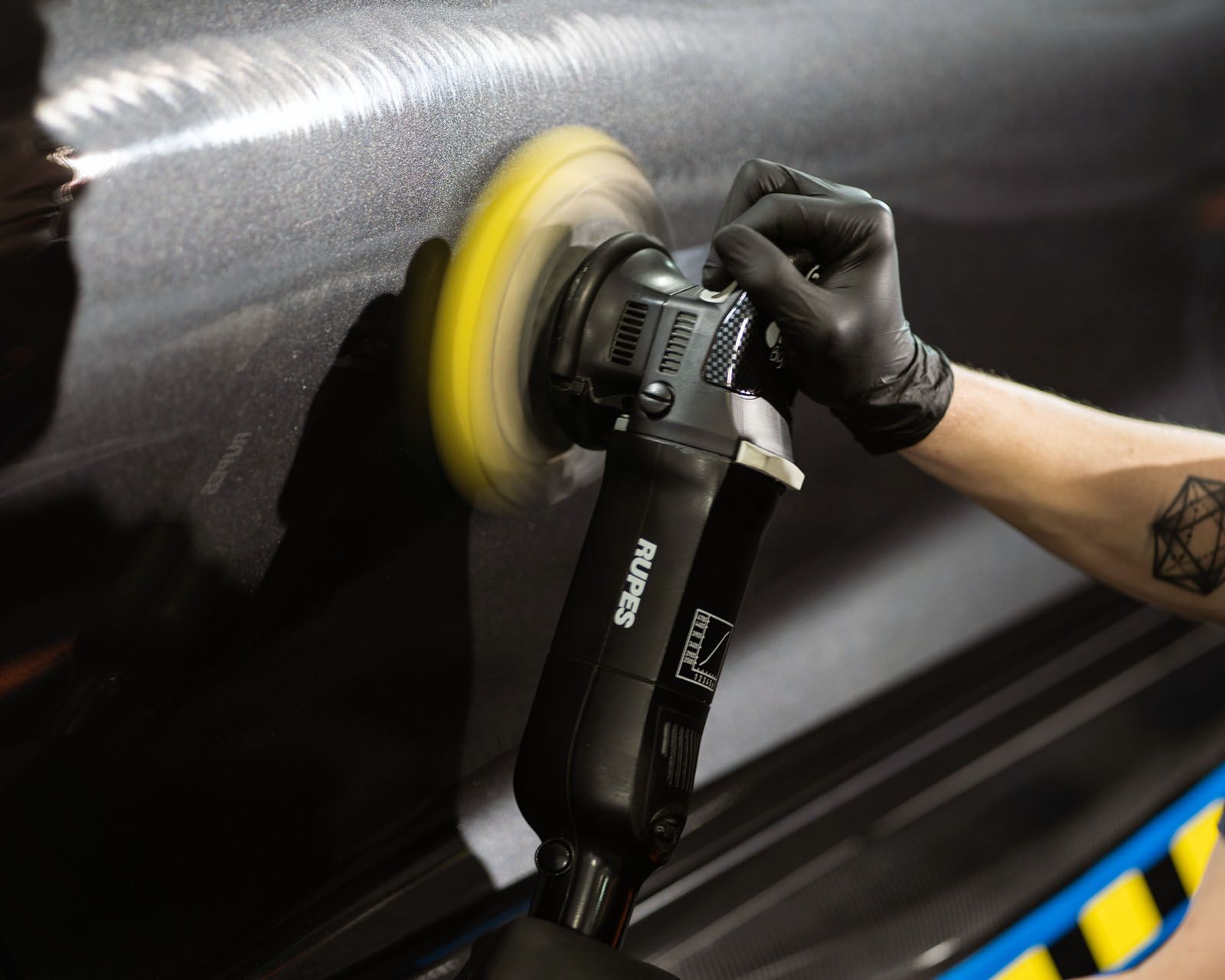 A person wearing black gloves using a polishing machine on a car's dark surface.