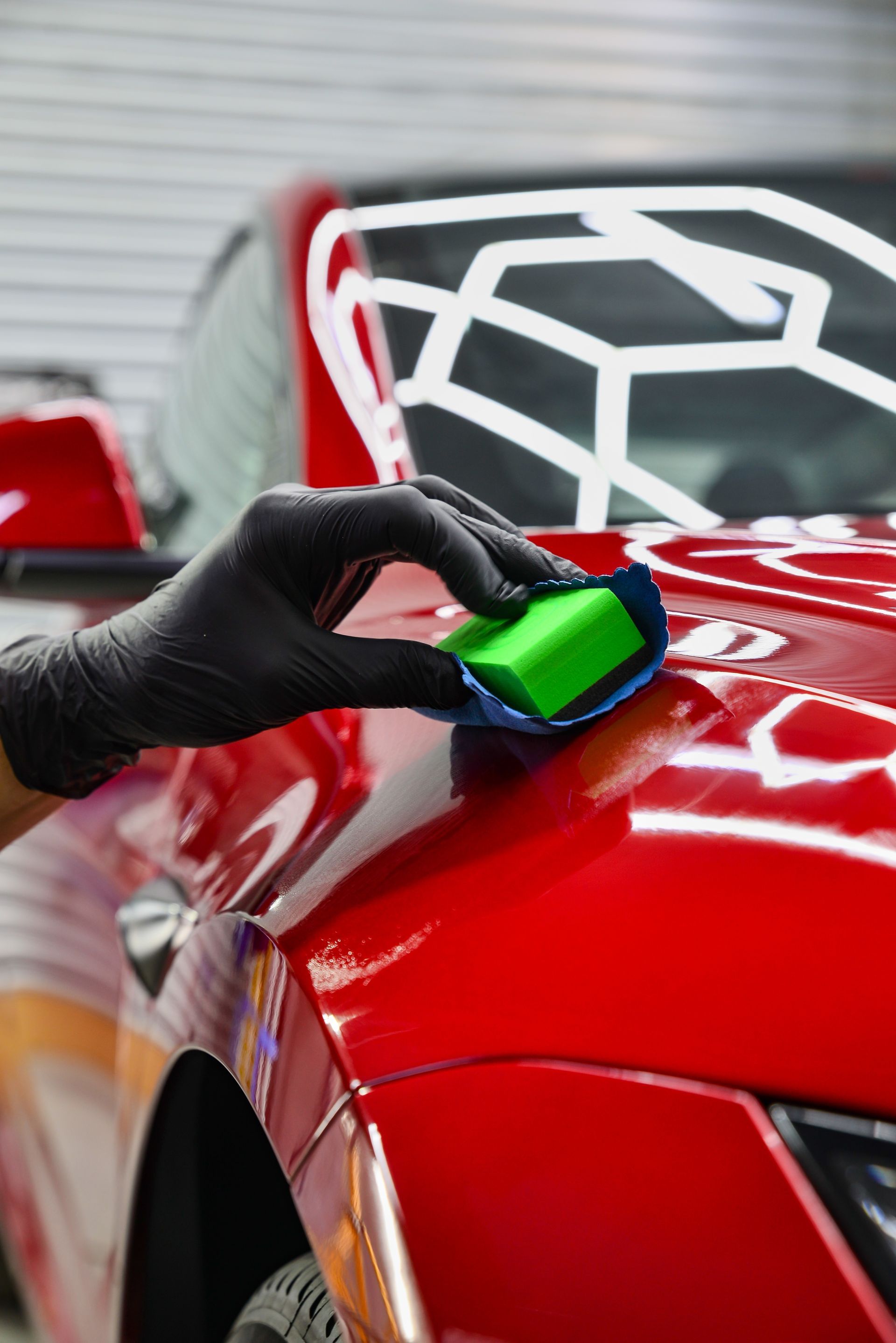 Black-gloved hand applying sealant to the red hood of a car with a green applicator.