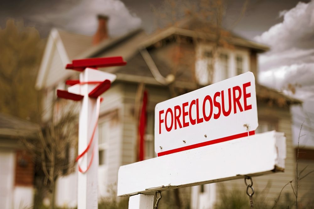 A close-up of a white foreclosure sign in the foreground, with a blurred residential house in the background.