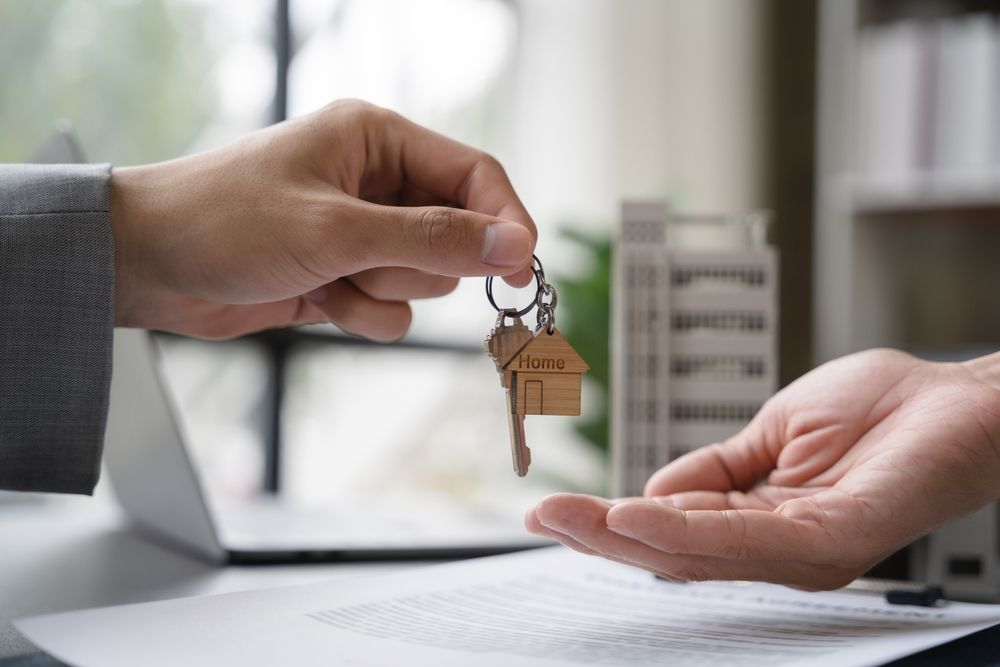 A hand passing keys with a small house-shaped keychain to another hand, set against an office desk with paperwork.