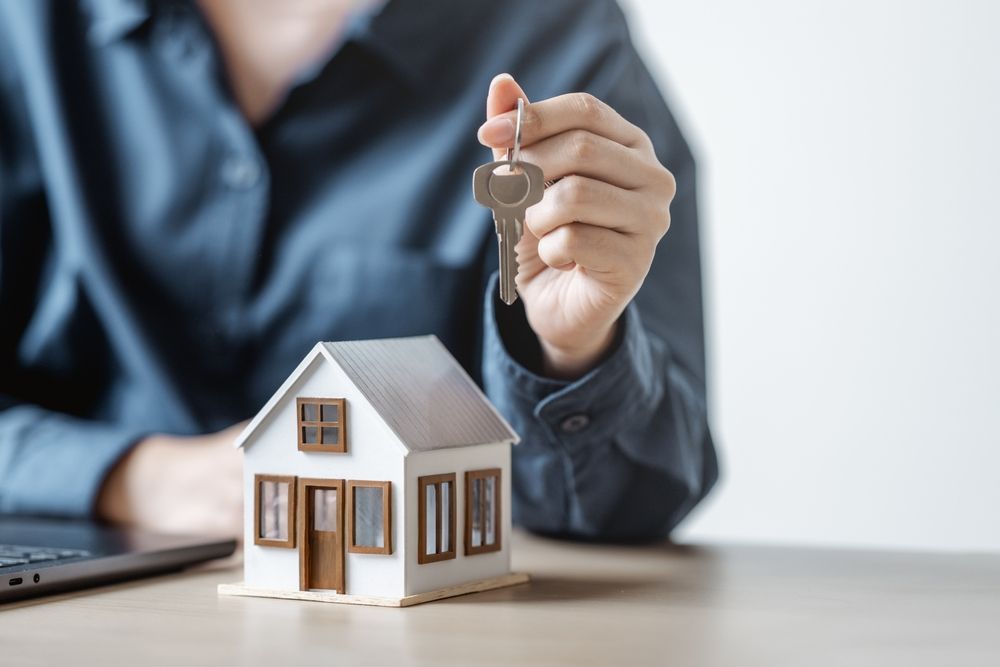 A person holding a key over a small model house on a desk.