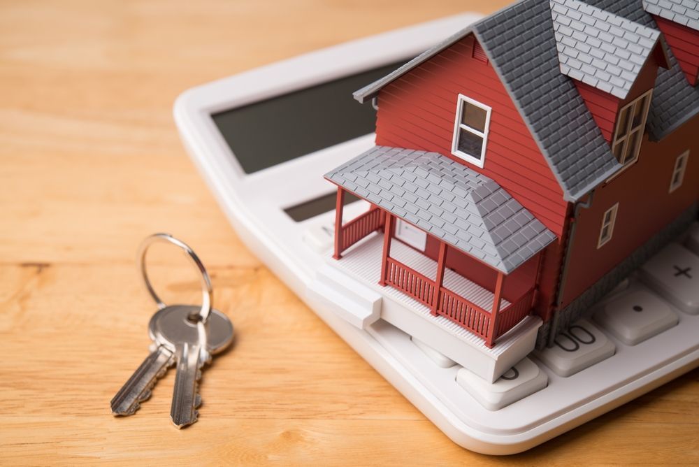 A model house sits on a white calculator next to a set of metal keys on a wooden table.