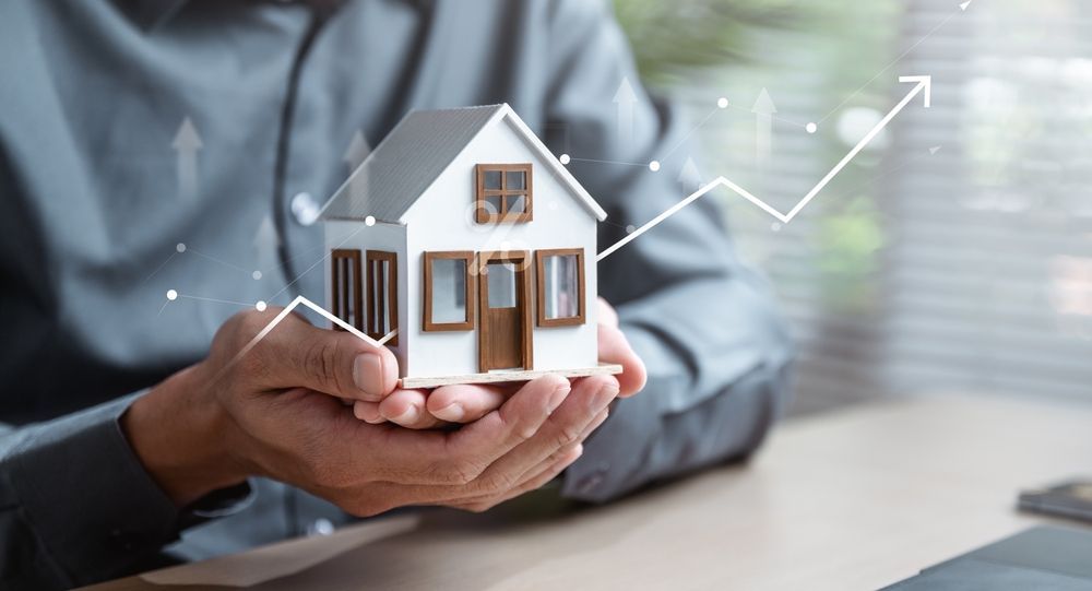 Hands holding a small model house with a digital line graph overlay showing an upward trend.