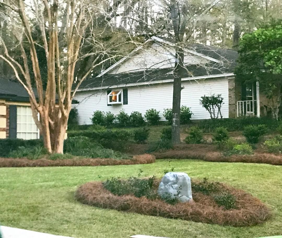 Suburban house with white siding, trees, shrubs, and a landscaped front yard with a stone feature.