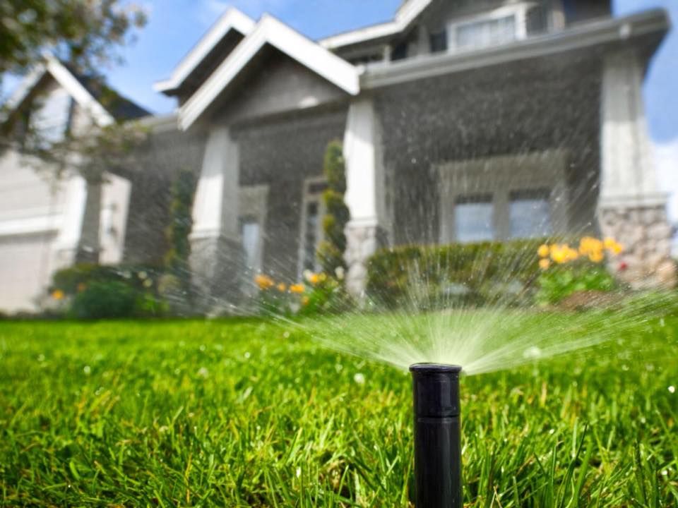 Sprinkler watering a green lawn in front of a gray house with flowers.