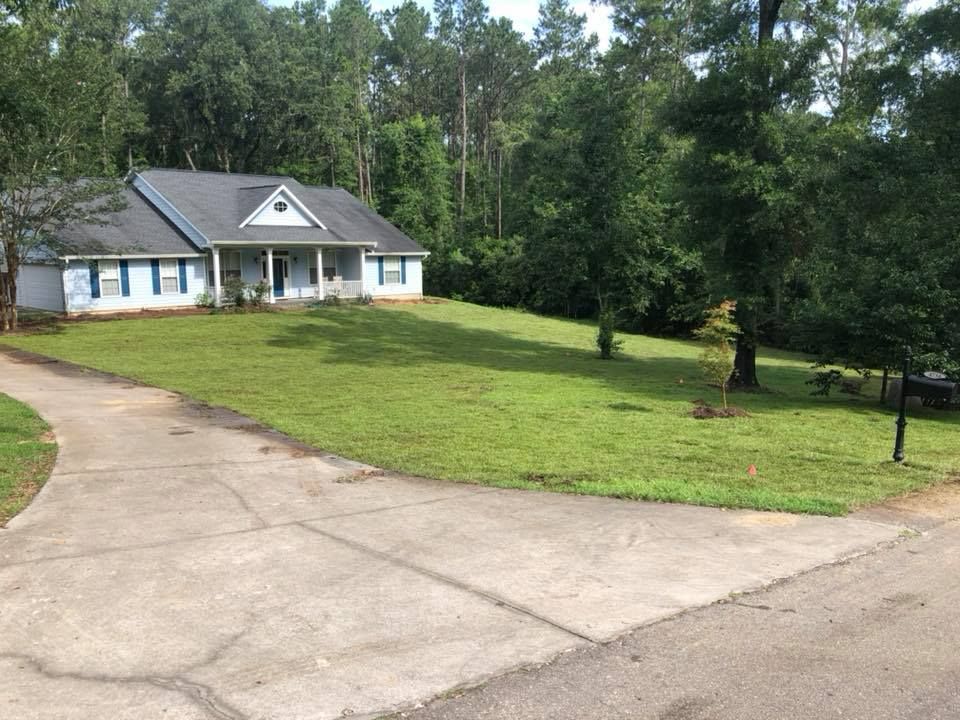 Single-story house with a gray roof, large green lawn, and curved driveway beside wooded trees.