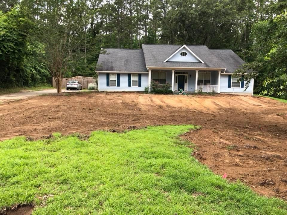 Single-story white house with blue shutters, front porch, and freshly graded brown yard in a wooded setting