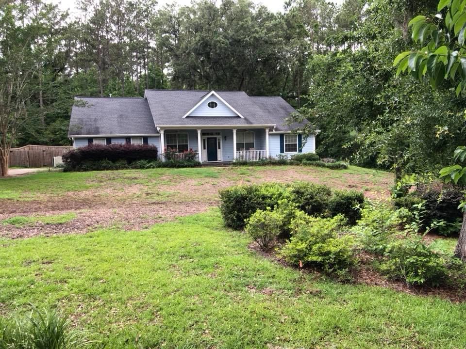 Single-story white house with a gray roof, front porch, and a large grassy yard surrounded by trees.