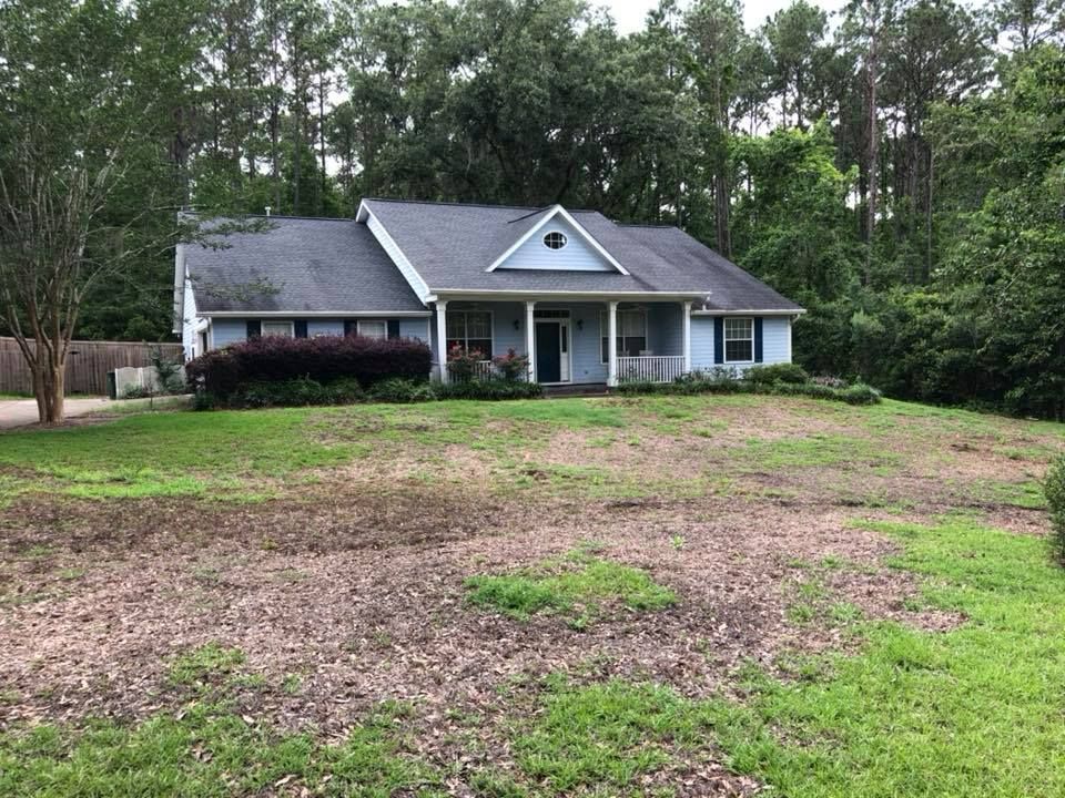Single-story gray house with a front porch, surrounded by trees and a patchy grassy yard
