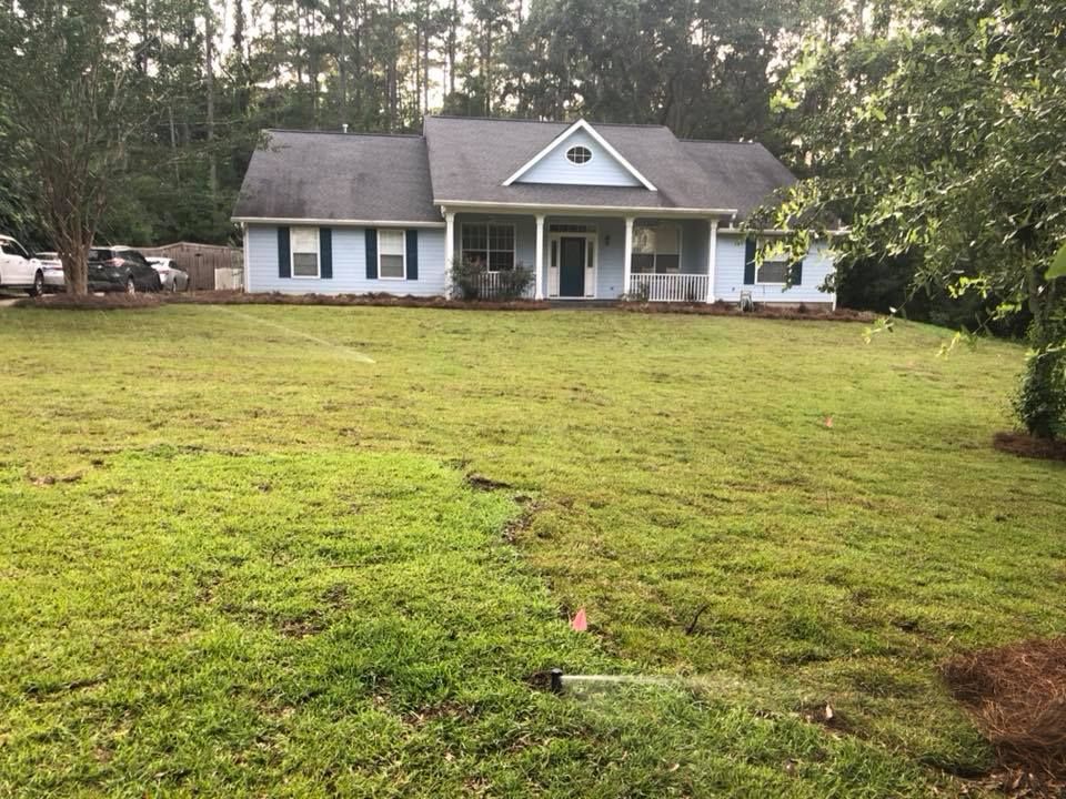 Single-story white house with a front porch, set behind a large grassy yard and trees.