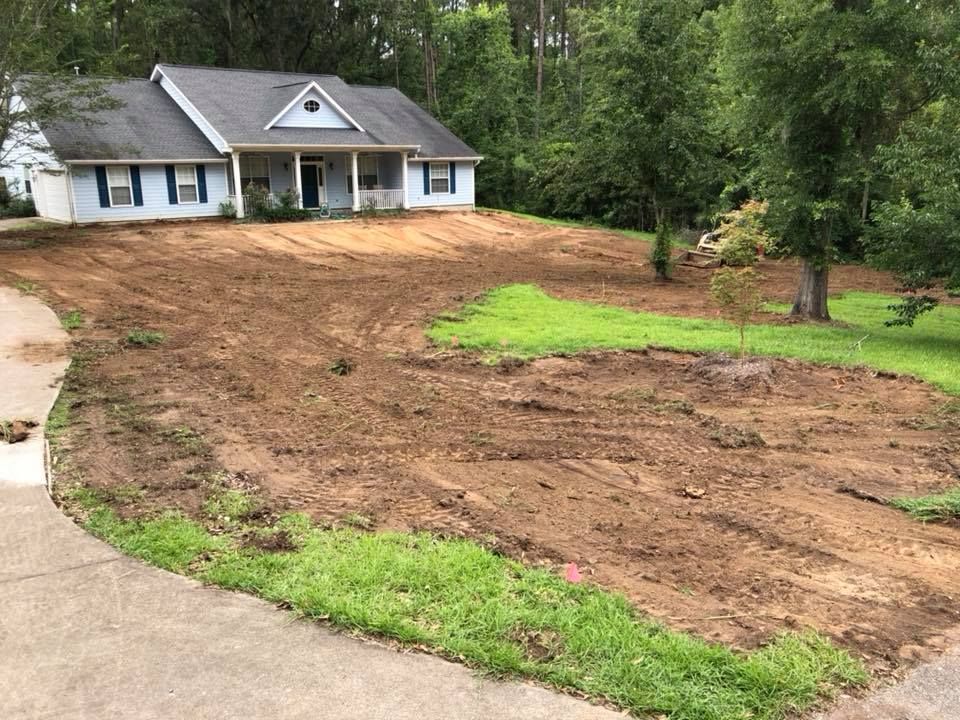 House on a wooded lot with a large dirt area and patches of grass beside a curved driveway