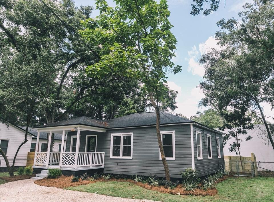 Gray house with white porch and tall trees in a leafy yard