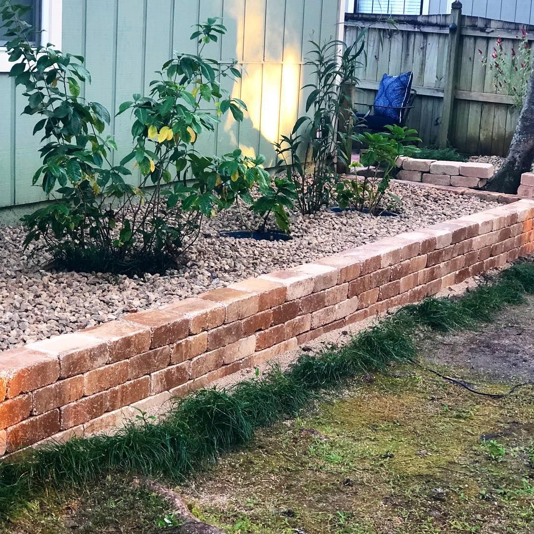 Raised brick garden bed with young plants and gravel beside a wooden fence in a yard