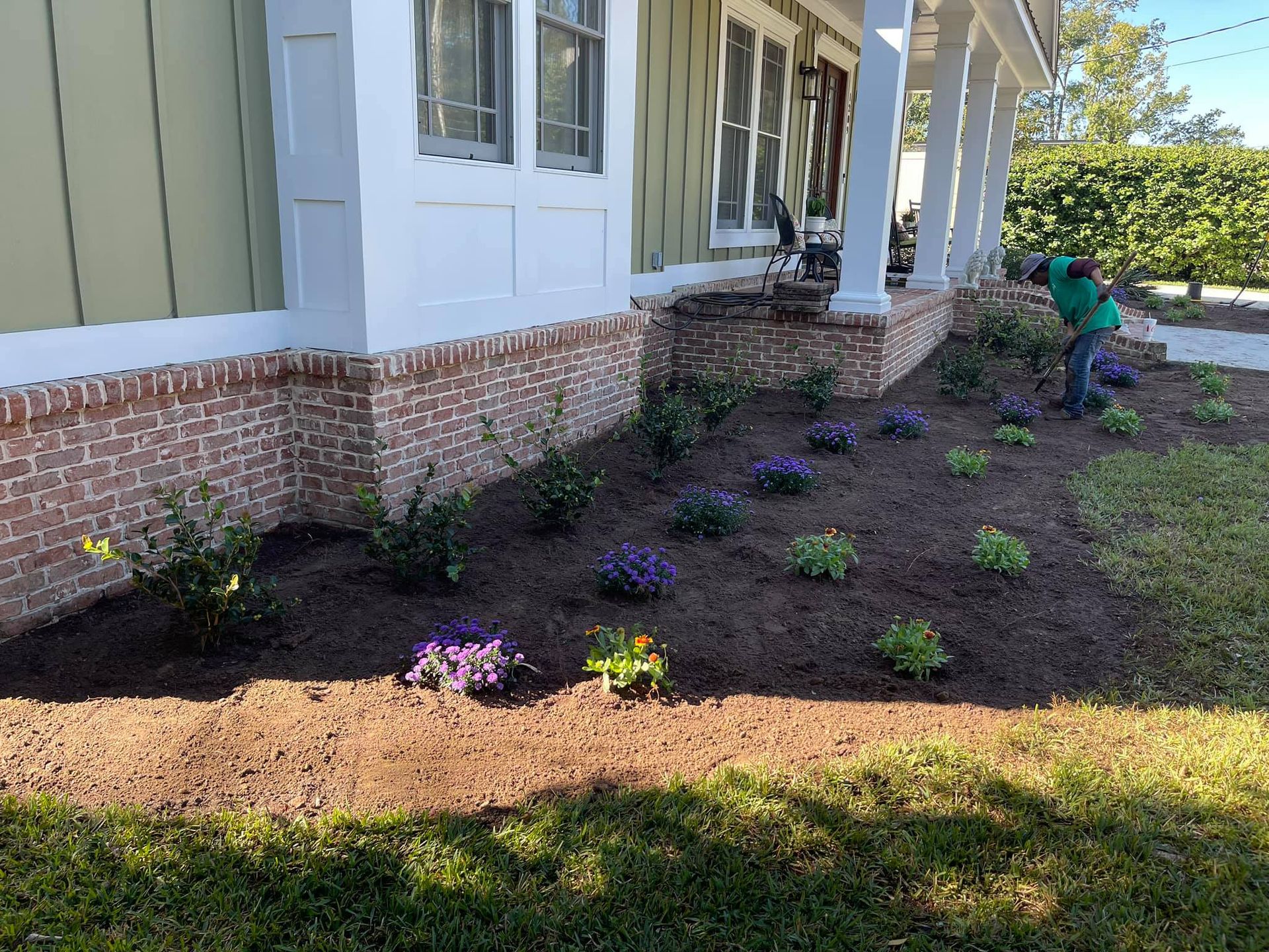 Landscaped flower bed beside a house with purple plants and a gardener using a leaf blower