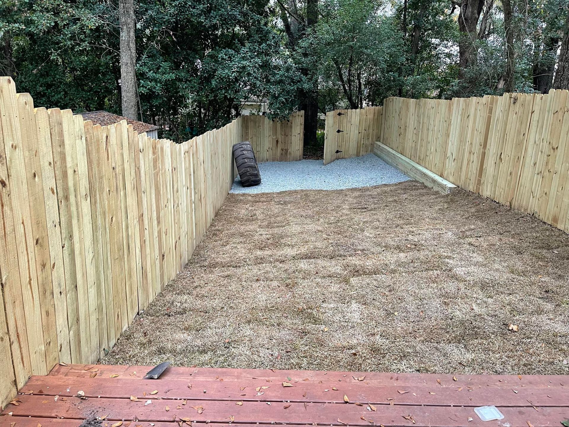New wooden fence enclosing a dirt yard with a gravel area at the far end and trees beyond.