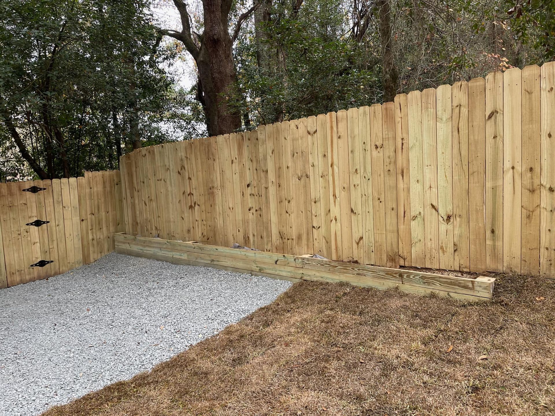 Gravel and dirt yard bordered by a tall wooden privacy fence and trees in the background