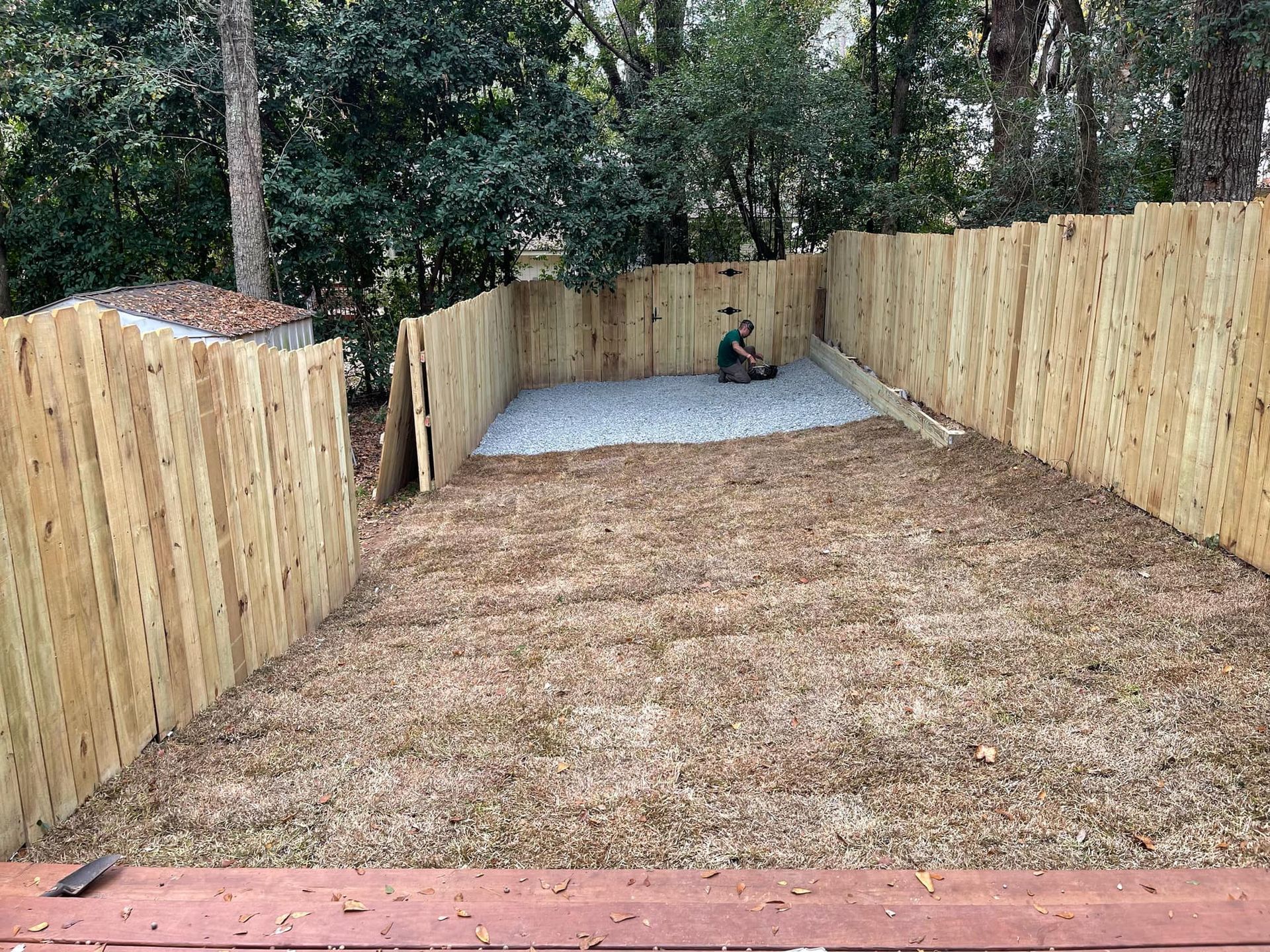 Wooden fenced backyard with a gravel pad and a small dog near the back wall.