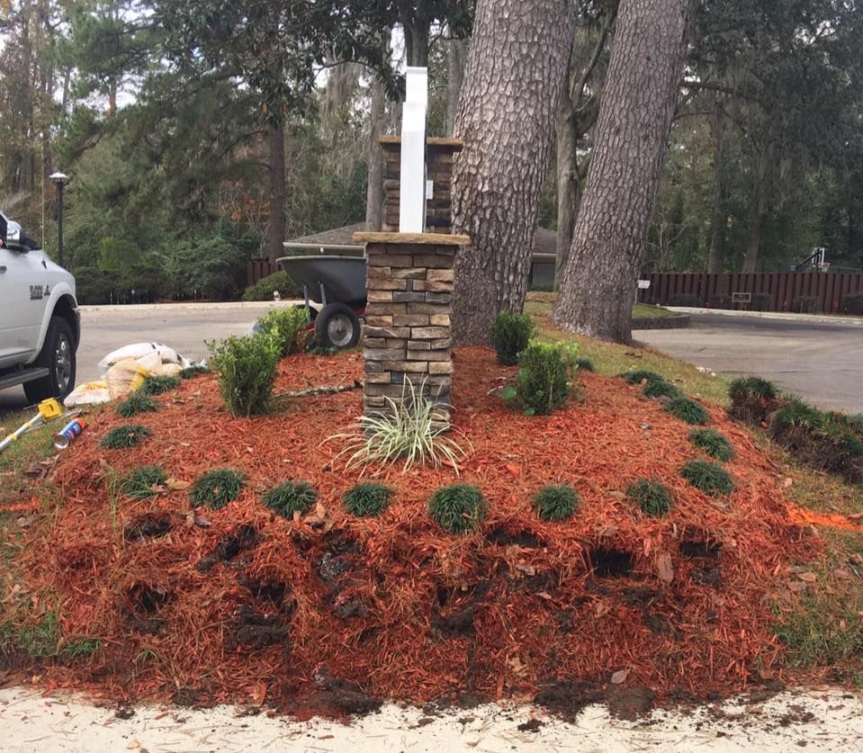Landscaped front yard with a stone pillar and white post among mulched shrubs and large trees