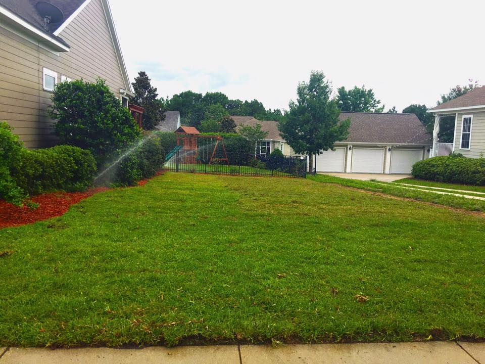 Suburban front yard with green lawn, brick houses, trees, and a driveway on a cloudy day