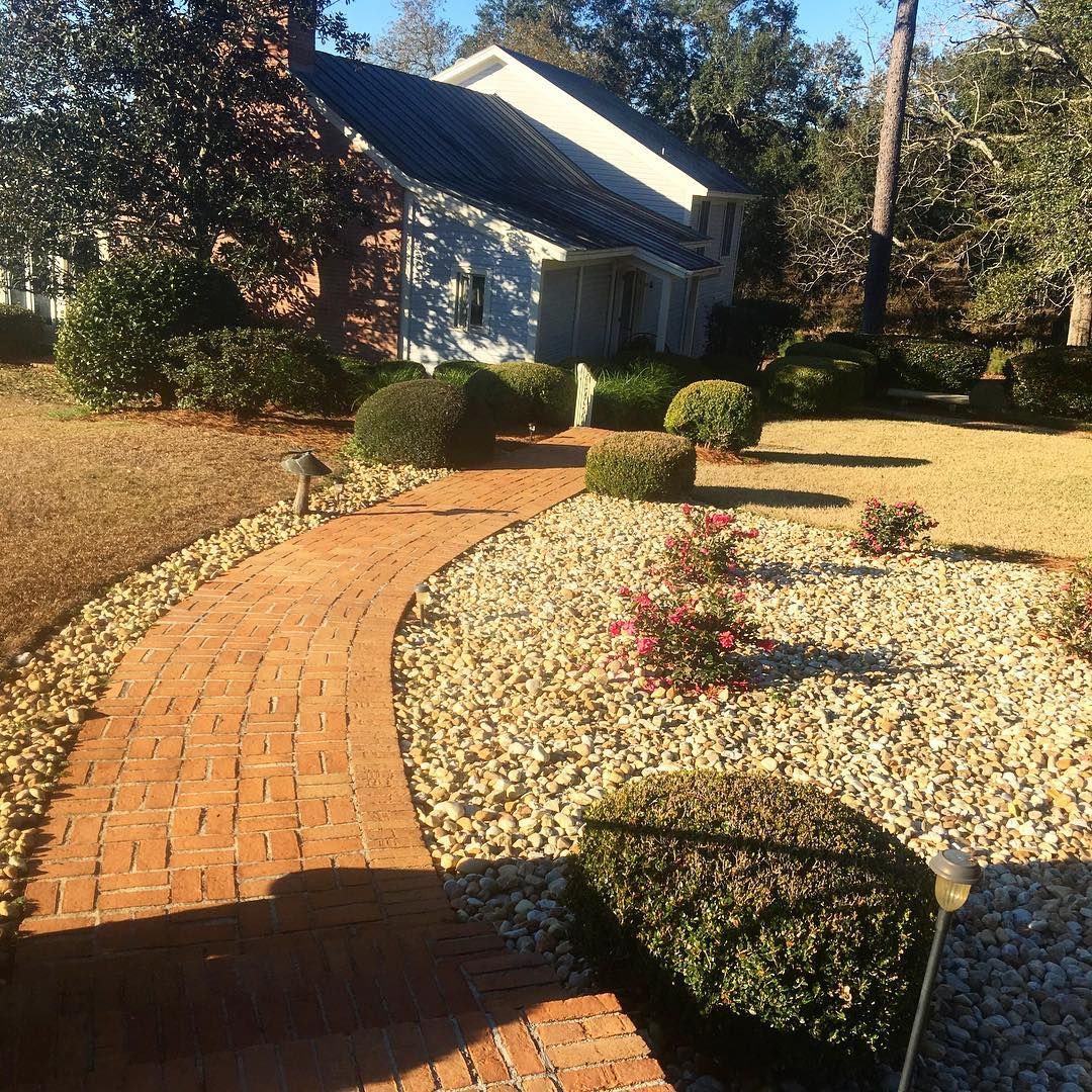Curved brick walkway through a landscaped yard with trimmed bushes and a house in the background
