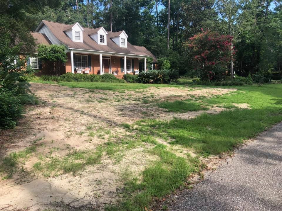 Suburban house with a front porch, dirt driveway, and grassy yard under trees