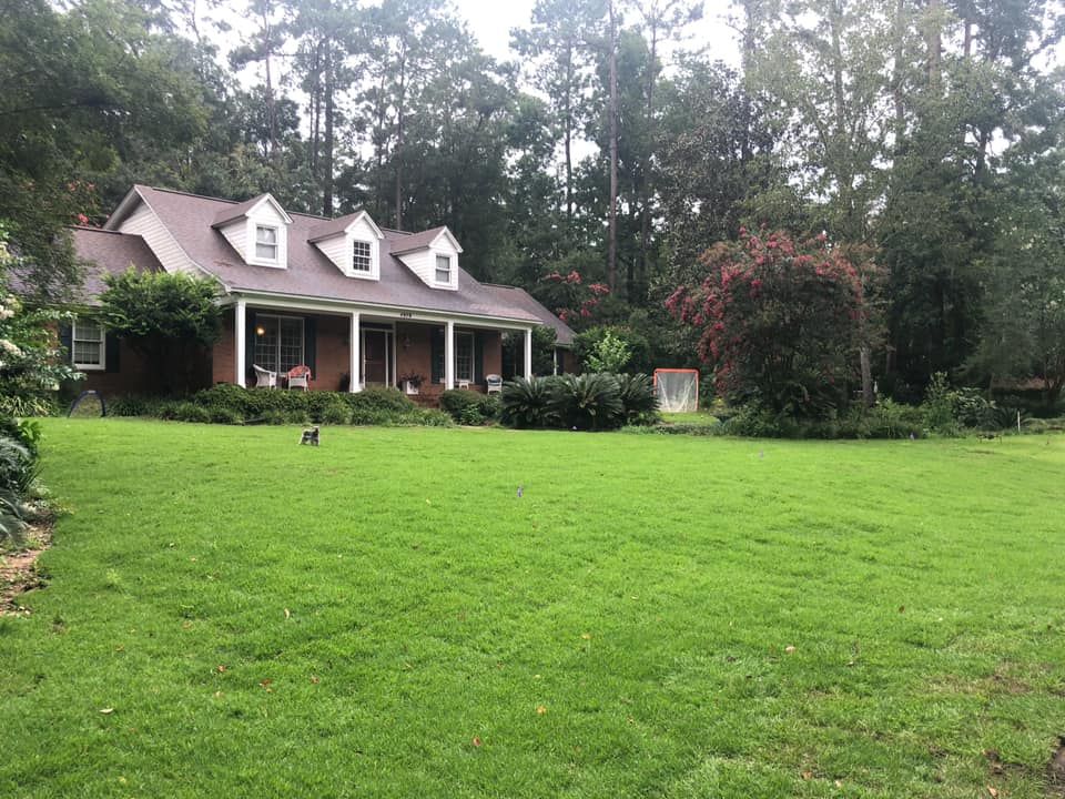 Suburban house with a brown roof, front porch, and a large green lawn bordered by trees