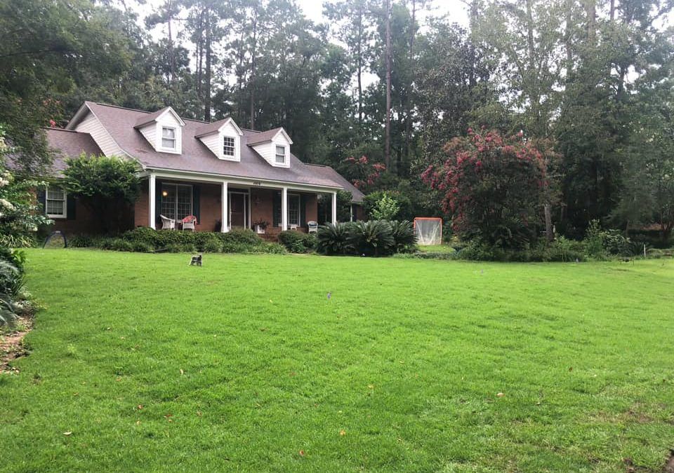 Ranch-style house with a front porch beside a large green lawn and trees