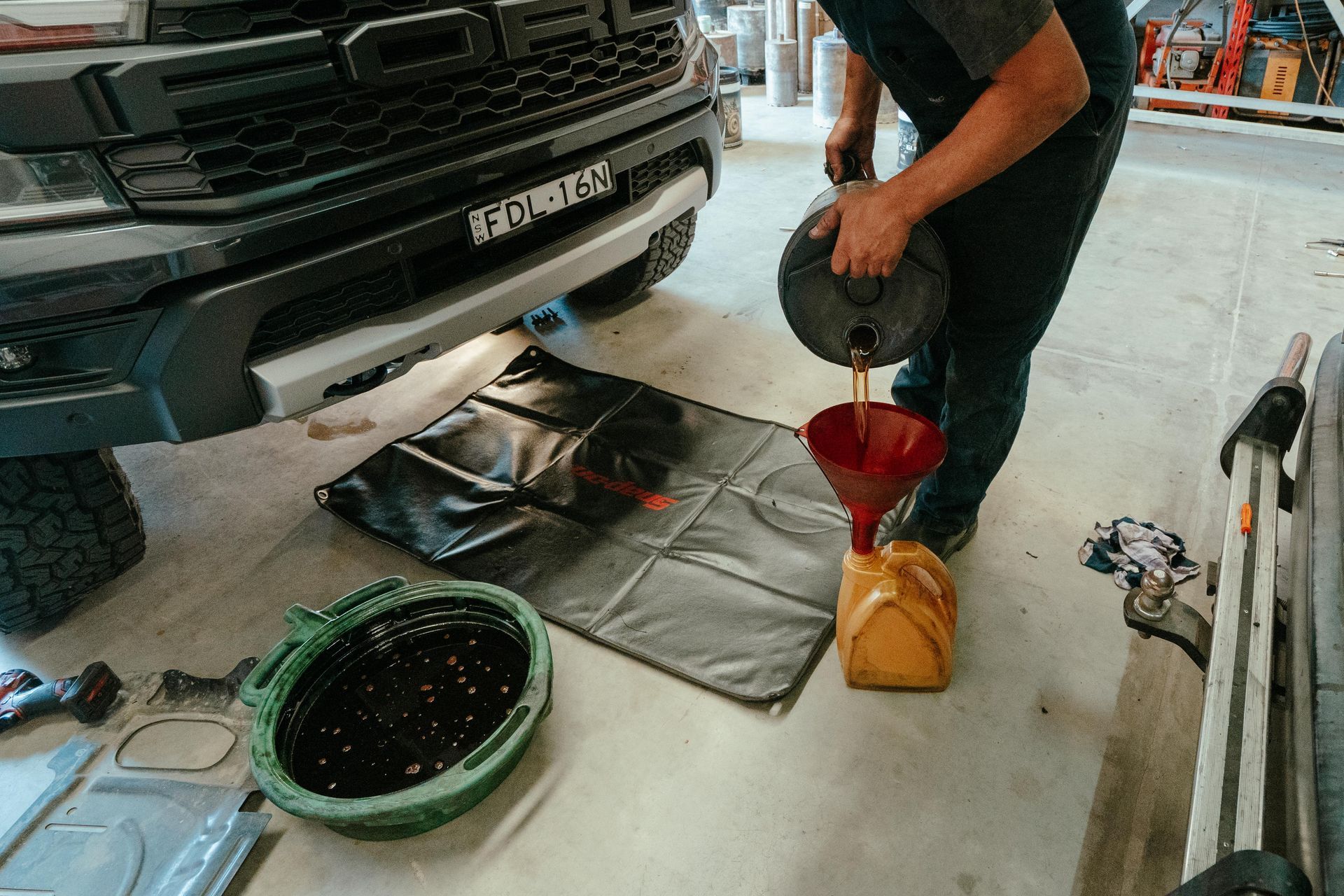 Person pouring oil from a container into a funnel leading to a yellow jug while changing oil under a truck. — Navan Mobile Mechanic in Corrimal, NSW