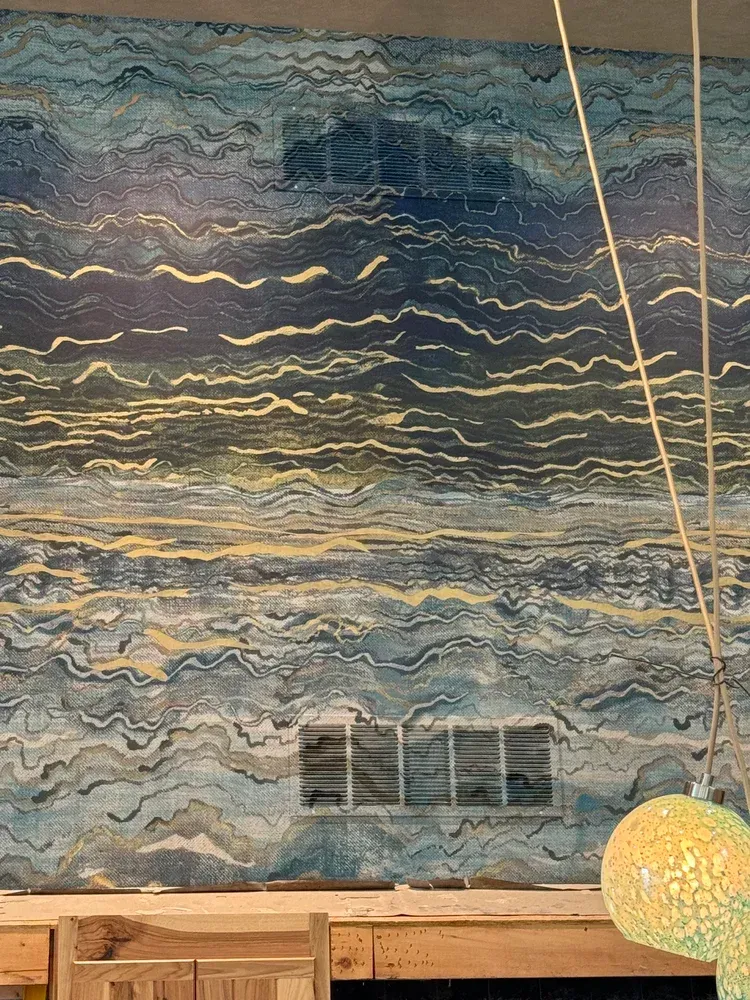 A wall mural featuring abstract, horizontal wavy lines in deep blue, gray, and gold, positioned above a wooden desk.