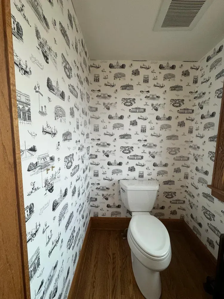 A toilet centered in a small bathroom with wood-look flooring and white wallpaper featuring black illustrations of buildings.