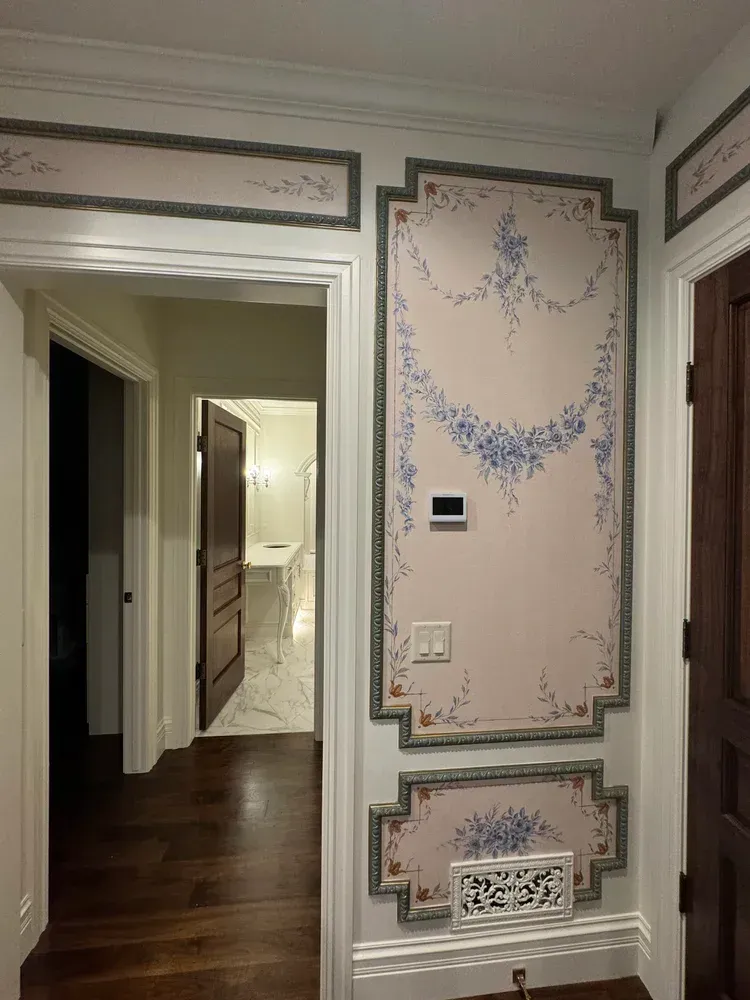 A hallway wall featuring panels with hand-painted blue floral garlands in a pale pink frame, beside a doorway.