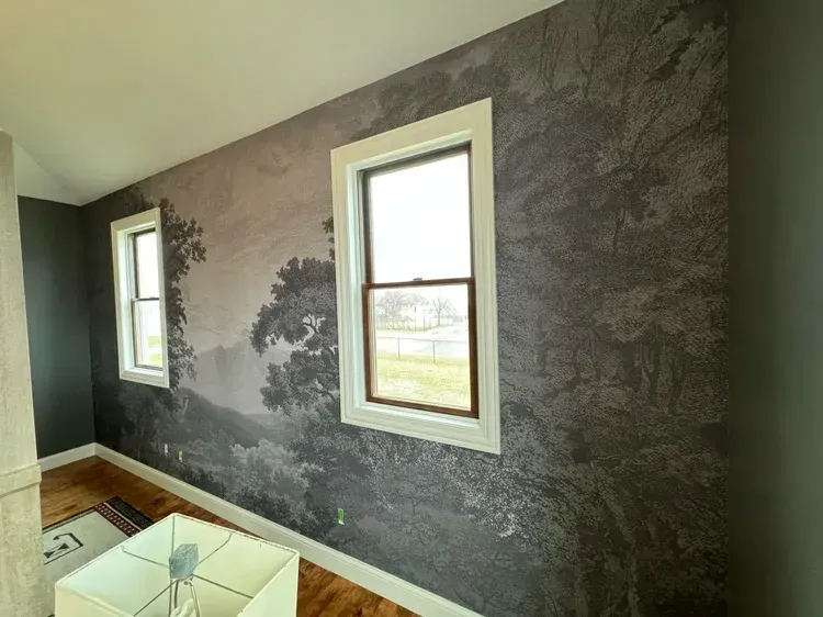 An interior room featuring a wall with a large-scale, monochromatic landscape mural of trees, set behind two white windows.