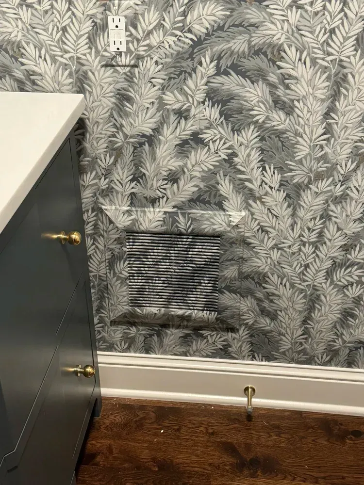 A wall vent cover disguised with matching wallpaper sits above dark hardwood floors near a cabinet.