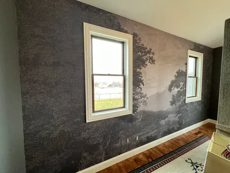 A wall with landscape-themed wallpaper showing trees and hills, featuring two white-framed windows looking outdoors.