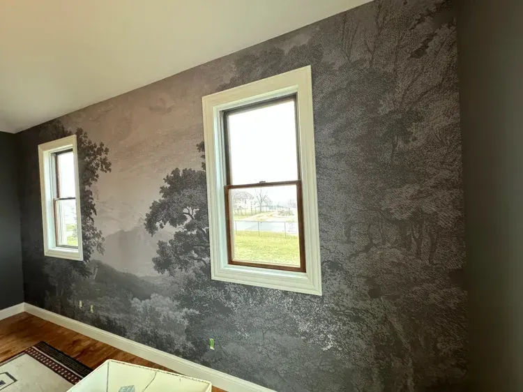 A wall covered in monochromatic scenic wallpaper featuring a landscape of trees and hills, framed by two white windows.