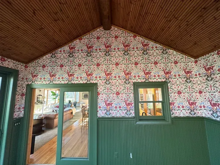 A room with green beadboard wainscoting, floral patterned wallpaper featuring red deer, and a wood-paneled peaked ceiling.