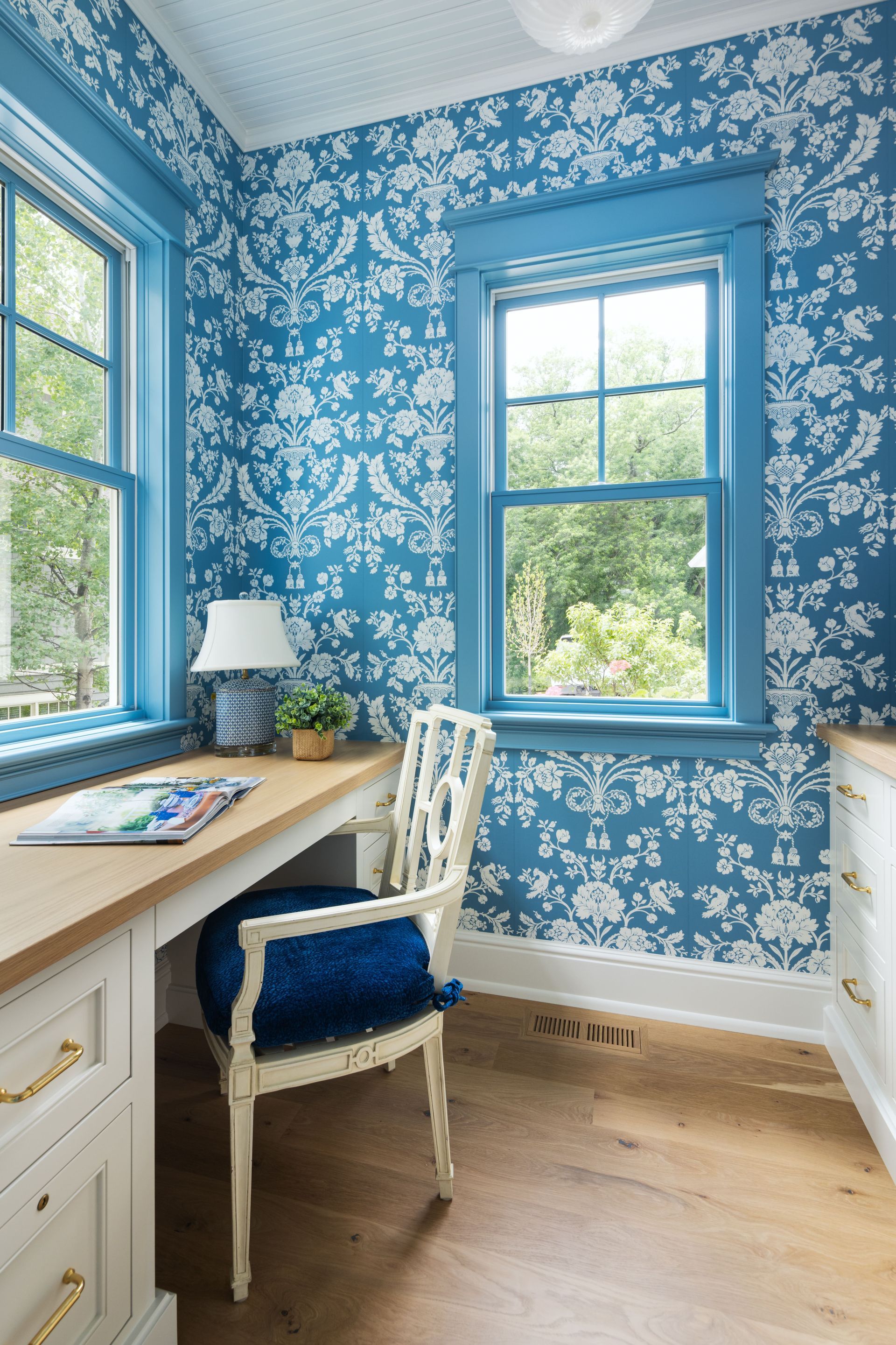 A bright home office corner with blue patterned wallpaper, a light wood desk, a blue cushioned chair, and two windows.