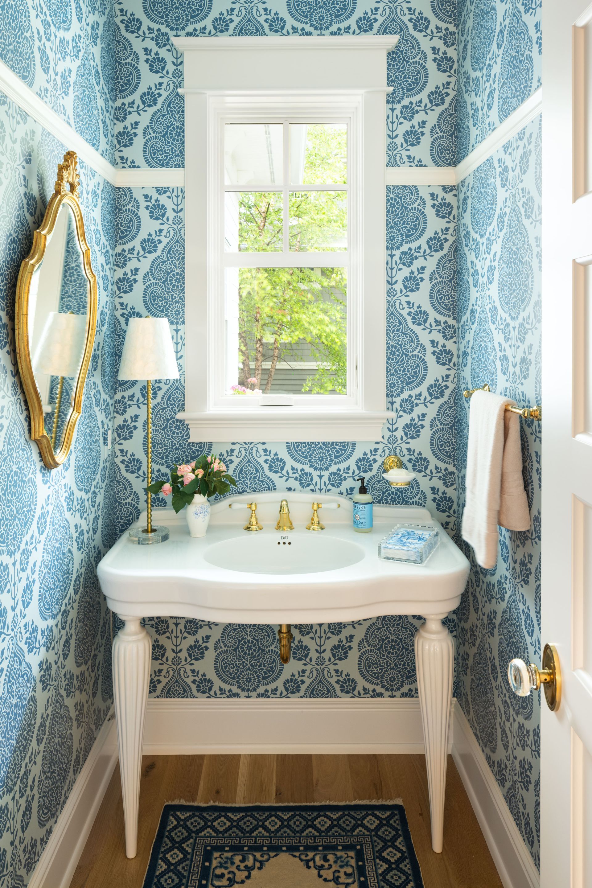 A small powder room with blue and white damask wallpaper, a pedestal sink, gold faucet, and an ornate gold wall mirror.