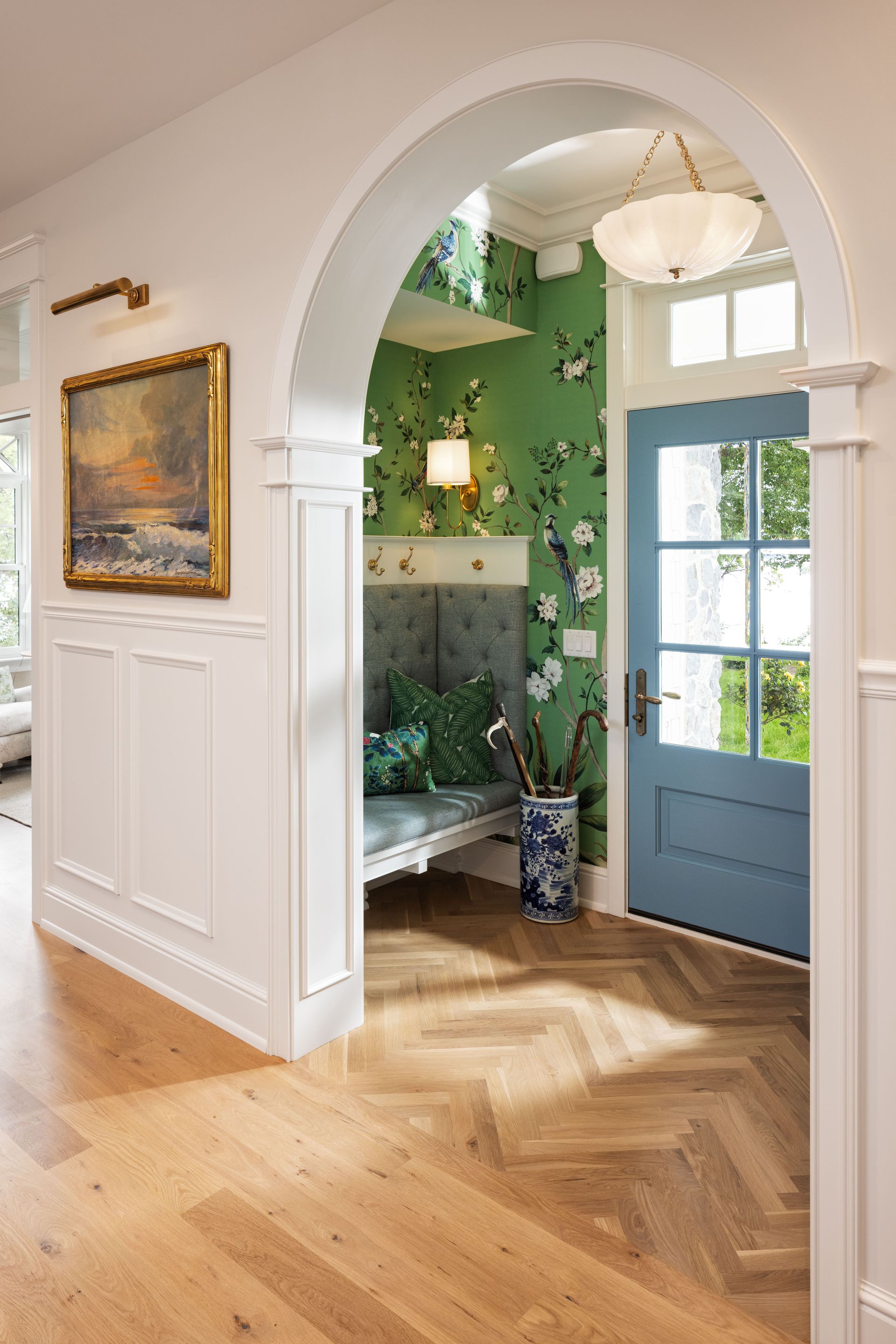 A white arched doorway reveals a blue front door, green patterned wallpaper, a gray built-in bench, and wood flooring.