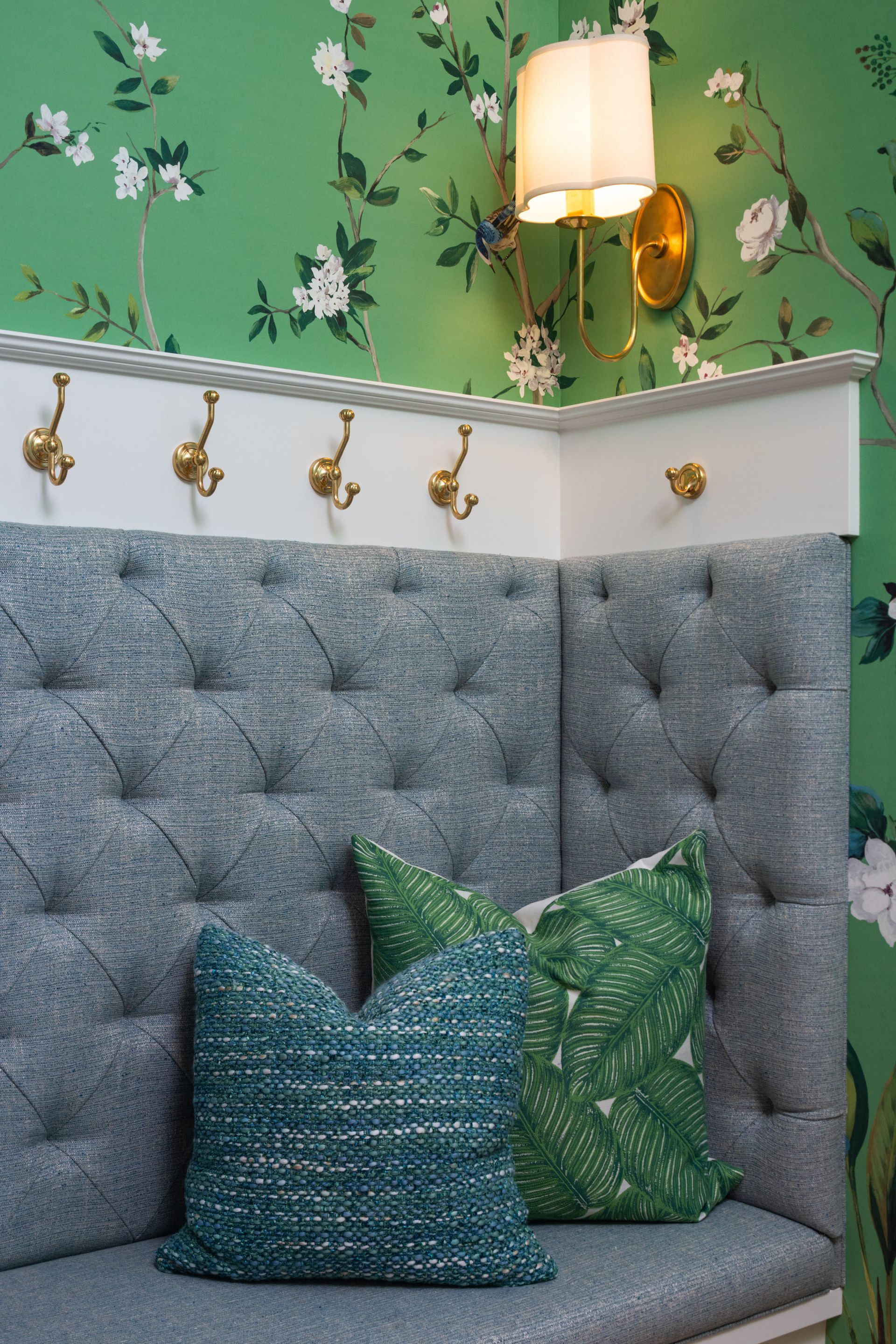 Gray tufted bench seat with two accent pillows, brass coat hooks on a white ledge, and green floral wallpaper with a light.