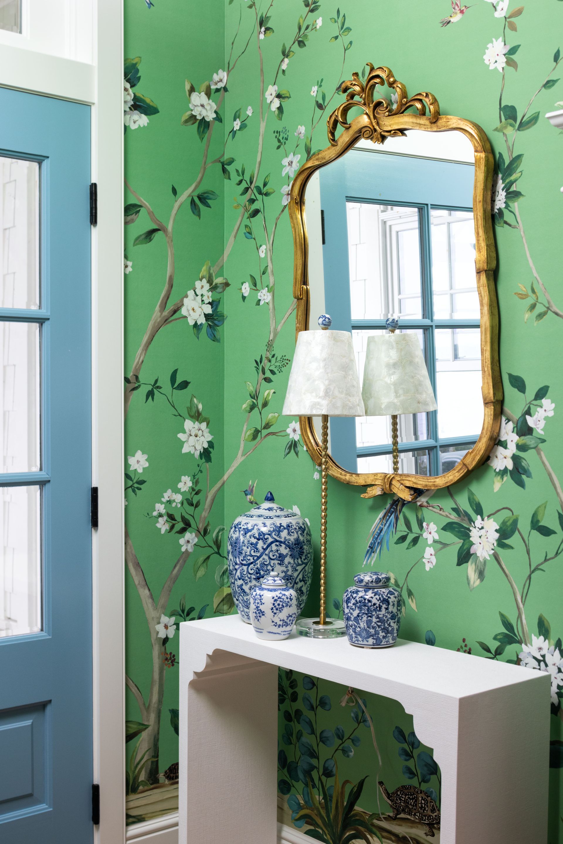 A white console table sits against green floral wallpaper, topped with two blue and white vases and a gold-framed mirror.