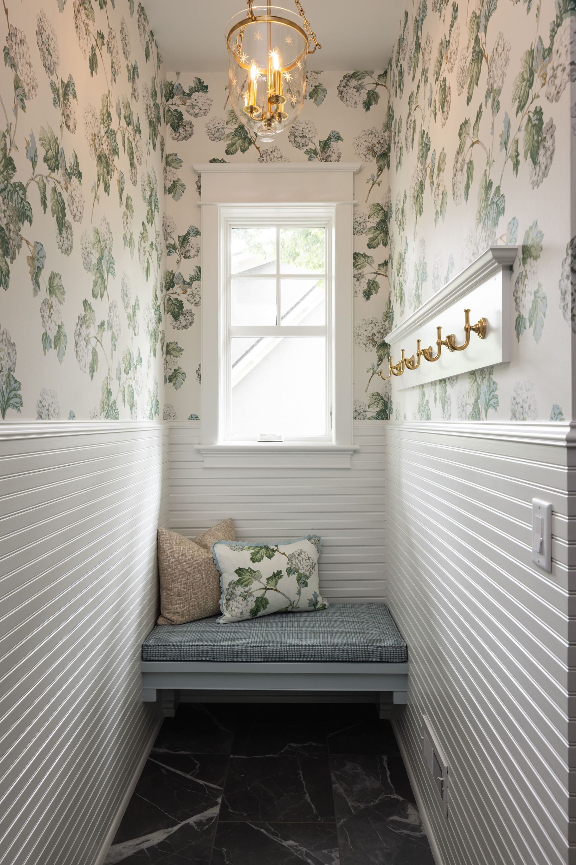 A narrow mudroom with green floral wallpaper, white beadboard, a window, a cushioned bench, and gold wall hooks.