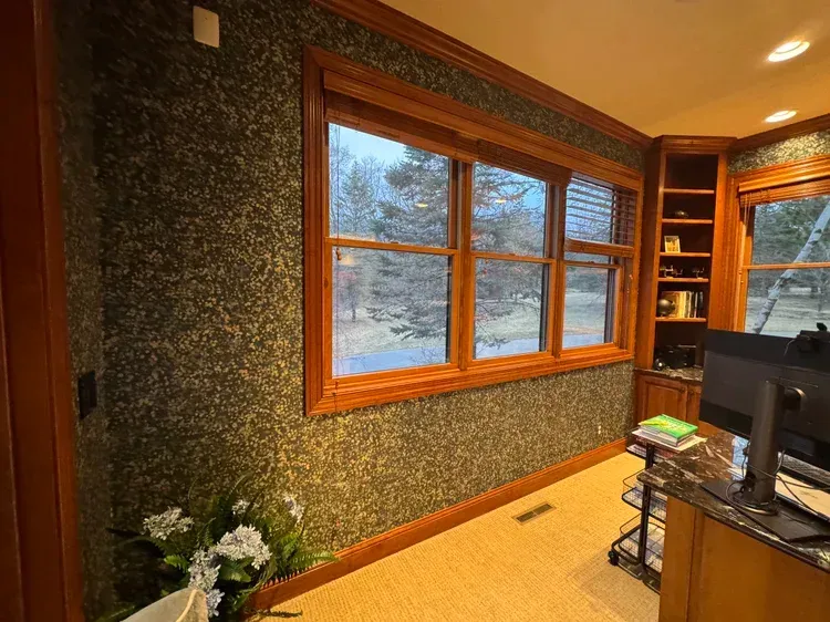A home office with dark patterned wallpaper, wood-trimmed windows, built-in shelving, and a computer desk.