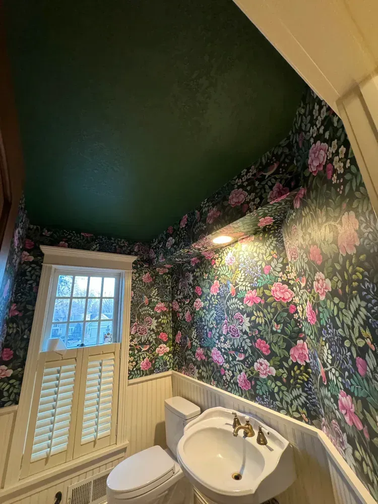 A small bathroom with dark floral wallpaper, a green ceiling, a white pedestal sink, and a window with white shutters.