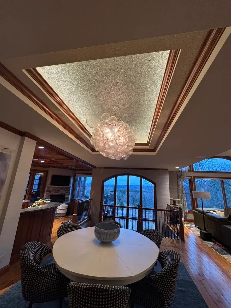 A bright dining area featuring a round white table, patterned chairs, and an ornate, glowing chandelier under a tray ceiling.
