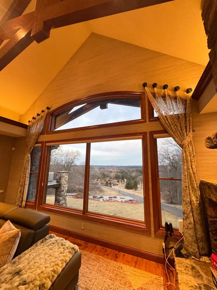 A large, wood-framed arched window in a vaulted room looks out over a rural landscape with trees and a road.