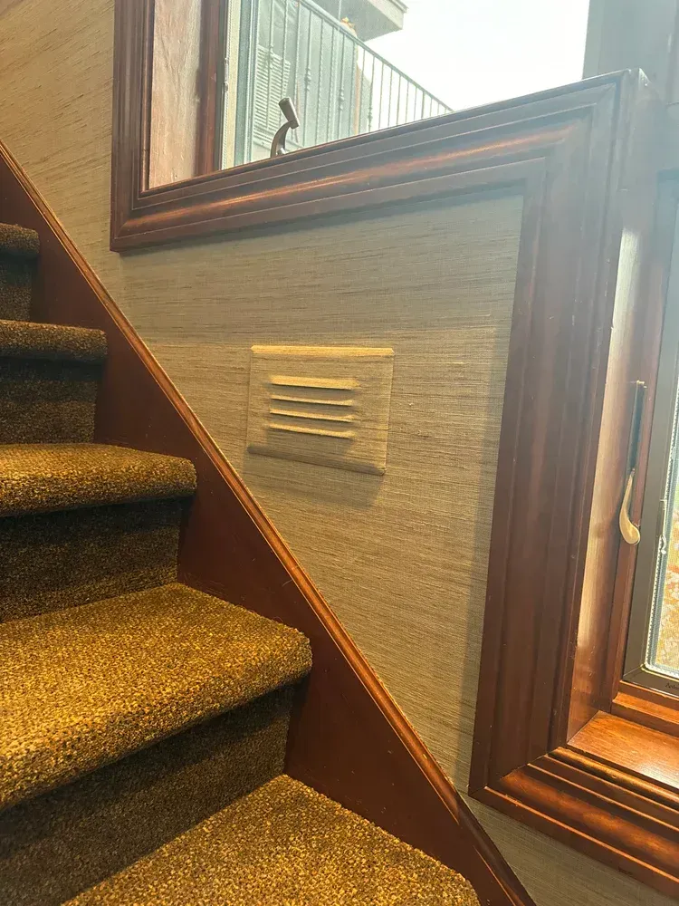 A beige louvered air vent mounted on a textured, grey-toned wall next to a wooden staircase and window frame.